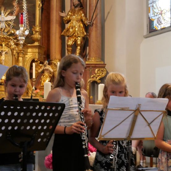 Drei junge Mädchen spielen Klarinetten in einer Kirche, mit einer goldenen Statue im Hintergrund.