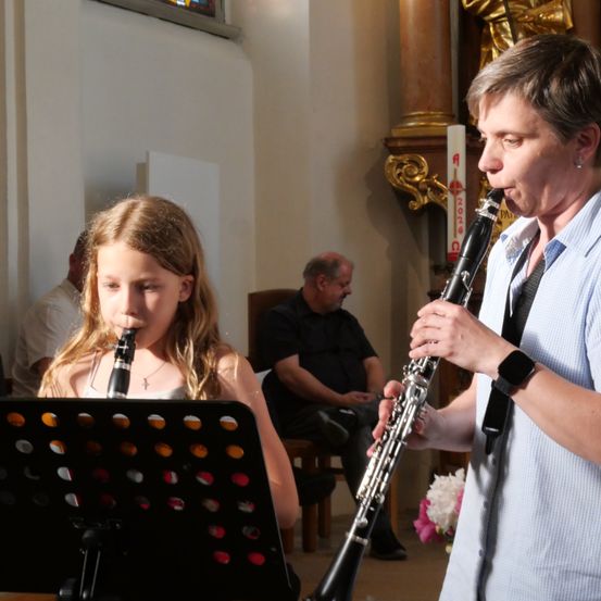 Ein junges Mädchen und eine erwachsene Frau spielen Klarinette in einer Kirche. Das Mädchen sitzt, und die Frau steht. Hinter ihnen sitzt ein Mann auf einem Stuhl.