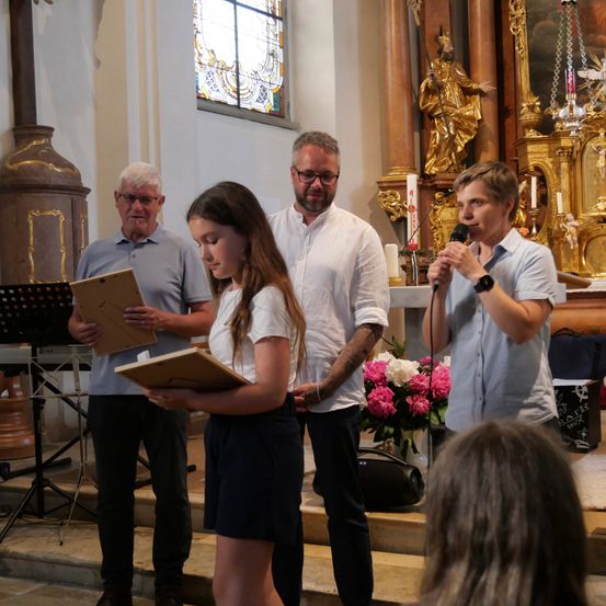 Eine Gruppe von Menschen steht in einer Kirche, ein Mann hält ein Mikrofon und eine Frau ein Buch. Hinter ihnen liest ein Mann aus einem Buch.