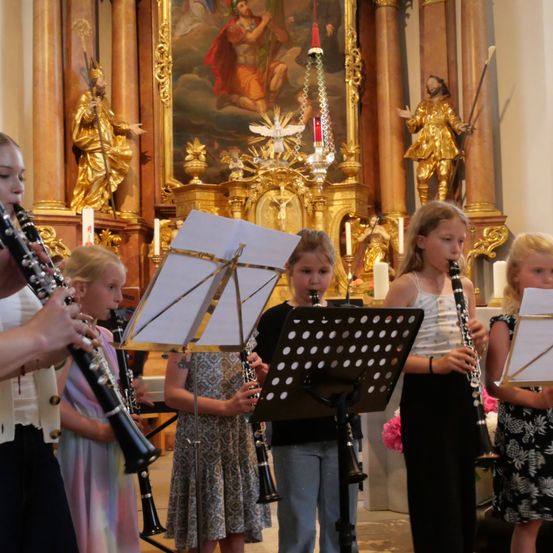 Fünf junge Mädchen spielen Klarinetten in einer Kirche. Sie stehen vor einem Notenständer mit Notenblättern. Dahinter befindet sich ein goldener Altar mit einem Gemälde und Statuen.