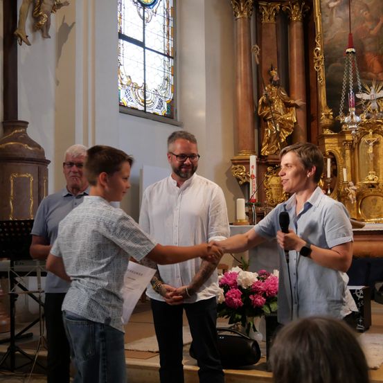 Ein Junge gibt einem Mann mit Mikrofon in einer Kirche die Hand. Hinter ihnen hält ein weiterer Mann ein Papier.