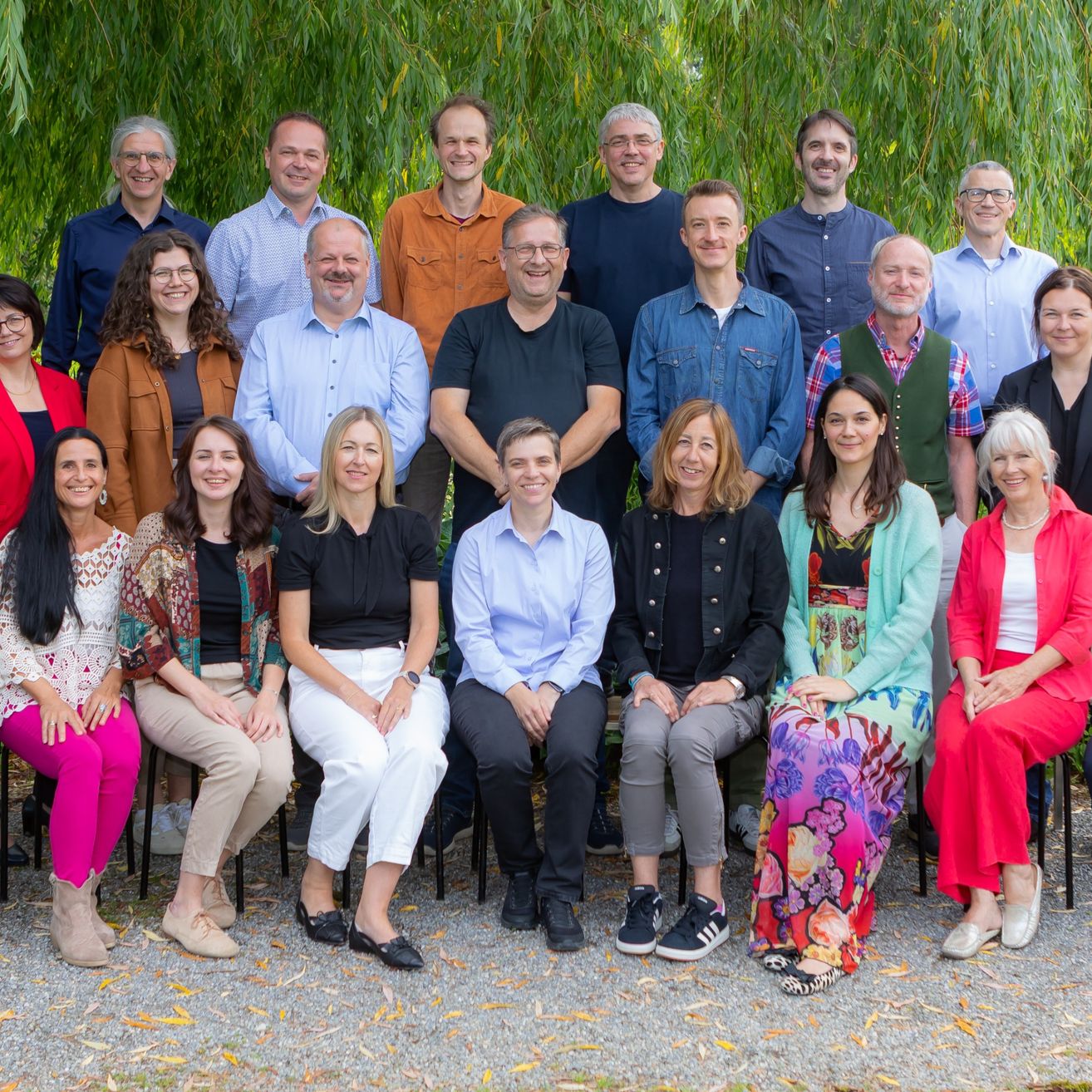Eine Gruppe von Menschen posiert für ein Foto in einer Außenumgebung mit Bäumen im Hintergrund. Sie sitzen auf Stühlen und stehen, in verschiedenen Outfits gekleidet.
