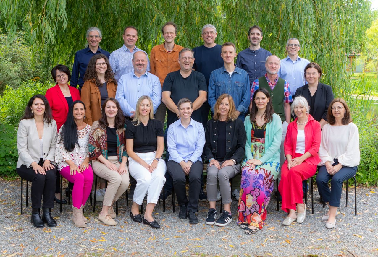 Eine Gruppe von Menschen posiert für ein Foto in einer Außenumgebung mit Bäumen im Hintergrund. Sie sitzen auf Stühlen und stehen, in verschiedenen Outfits gekleidet.