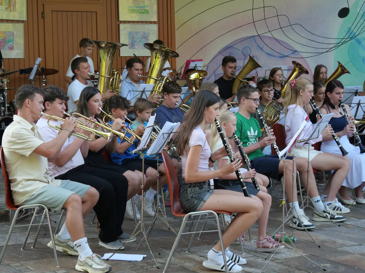 Eine Gruppe junger Musiker, Jungen und Mädchen, spielen verschiedene Instrumente wie Trompeten, Posaunen und Klarinetten in einem Raum. Sie sitzen auf Stühlen und scheinen für eine bevorstehende Aufführung zu proben.