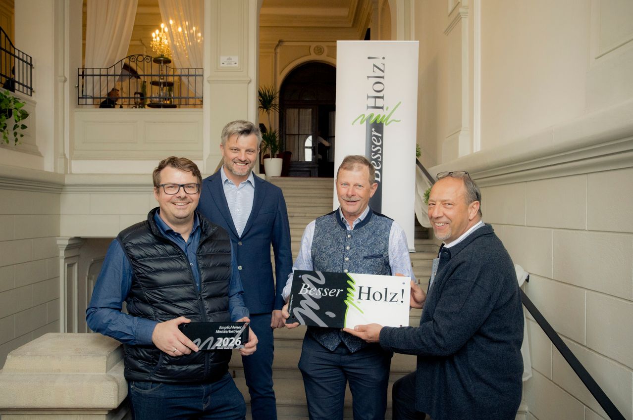 Four men stand in front of a staircase holding plaques. One man holds a sign that says Besser Holz. Behind them is a banner with the same text.
