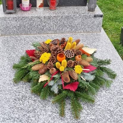 A wreath of pine cones, leaves, and flowers adorns a gray tombstone on a grassy field. Two lit candles and other small decorations are placed on the tombstone.