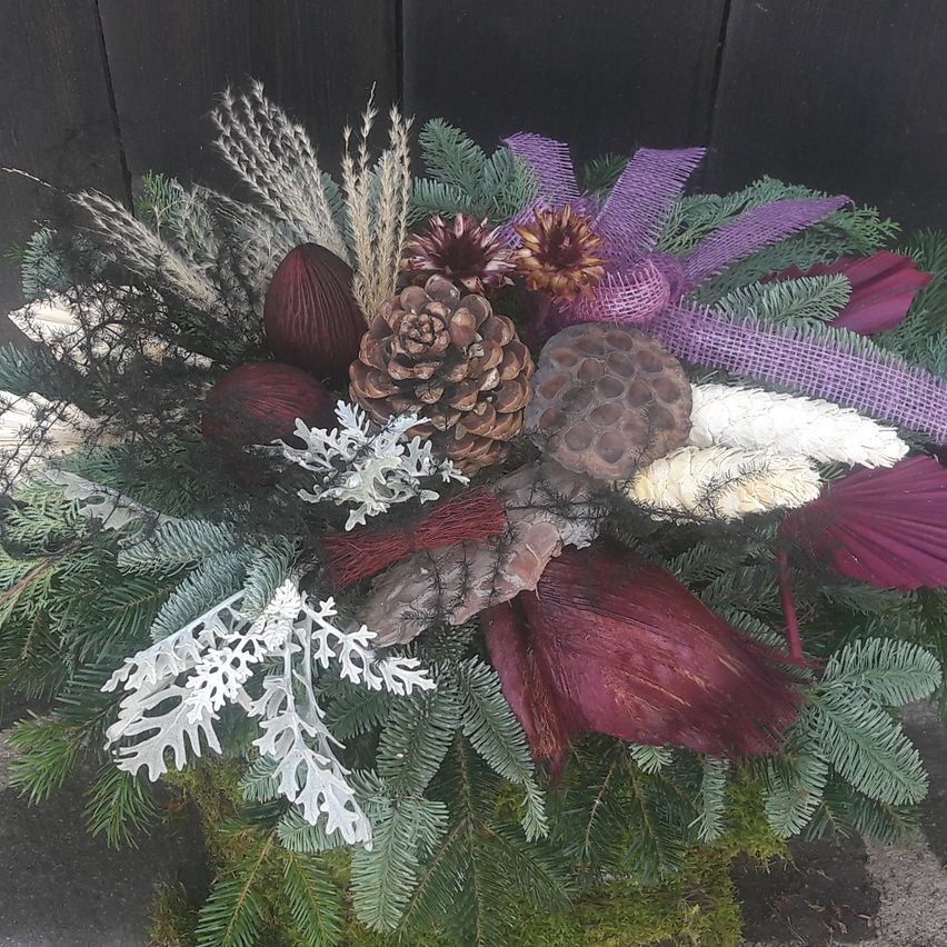Ein dekoratives Blumenarrangement mit getrockneten Blumen, Tannenzapfen und Grünpflanzen, akzentuiert mit lila Bändern und auf einer moosbedeckten Oberfläche platziert.