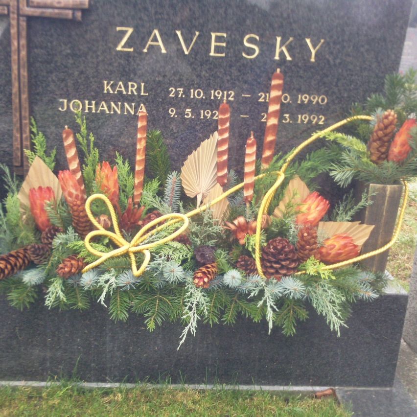 Ein Grabstein mit dem Namen Zavesky. Die Namen Karl und Johanna sind mit ihren Geburts- und Sterbedaten eingraviert. Der Grabstein ist mit Tannenzapfen, Kerzen und einer Schleife geschmückt.