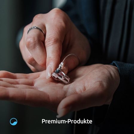 Eine Hand hält ein kleines elektronisches Gerät, wahrscheinlich ein Hörgerät, mit einem Kabel. Der Hand hat einen Ring am Finger. Das Bild hat einen Wasserzeichen mit den Worten 'Premium-Produkte'.