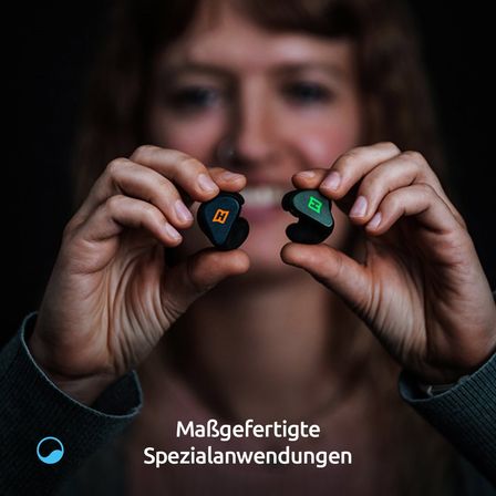 Eine Frau hält zwei schwarze Ohrhörer mit dem Buchstaben H und grünen Lichtern in der Hand und zeigt sie. Der Text lautet 'Maßgefertigte Spezialanwendungen.'