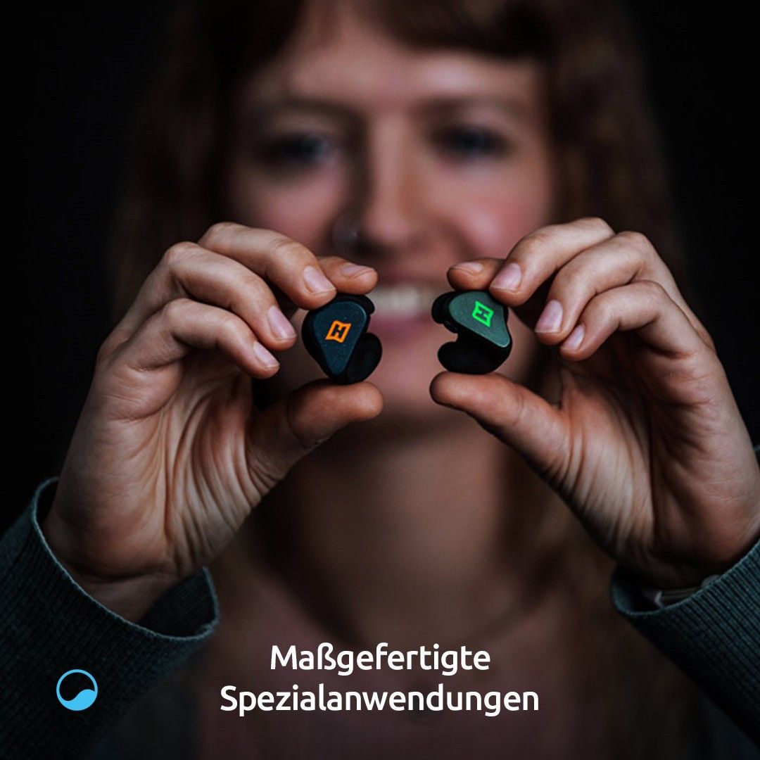 Eine Frau hält zwei schwarze Ohrhörer mit dem Buchstaben H und grünen Lichtern in der Hand und zeigt sie. Der Text lautet 'Maßgefertigte Spezialanwendungen.'