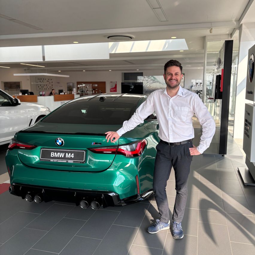 Ein Mann in einem weißen Hemd steht neben einem grünen BMW M4 Auto in einem Autohaus.
