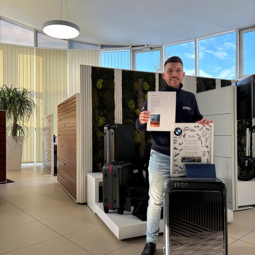 Ein Mann steht in einer Ausstellungshalle, lächelt und hält ein Zertifikat und ein Bild in der Hand. Im Hintergrund befinden sich große Fenster und eine moderne Inneneinrichtung mit Holzpaneelen und einem Topfplant.