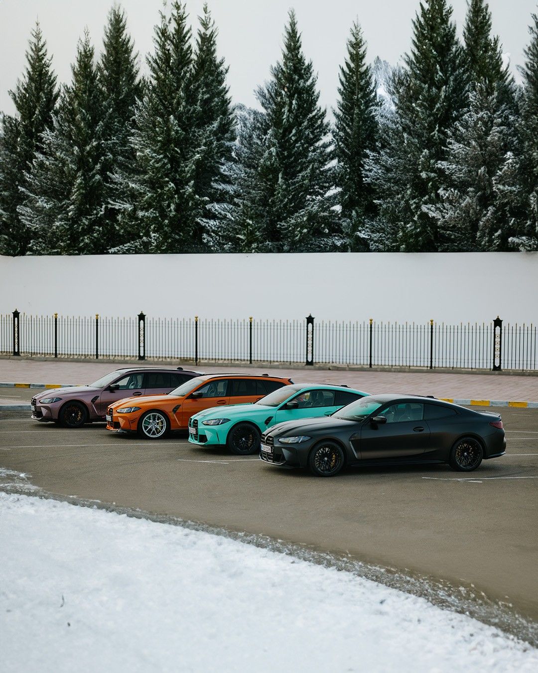 Ein verschneiter Parkplatz mit vier Autos in verschiedenen Farben, darunter ein schwarzes Auto, ein türkisfarbenes Auto, ein orangefarbenes Auto und ein lilafarbenes Auto, vor einer weißen Wand und Bäumen.