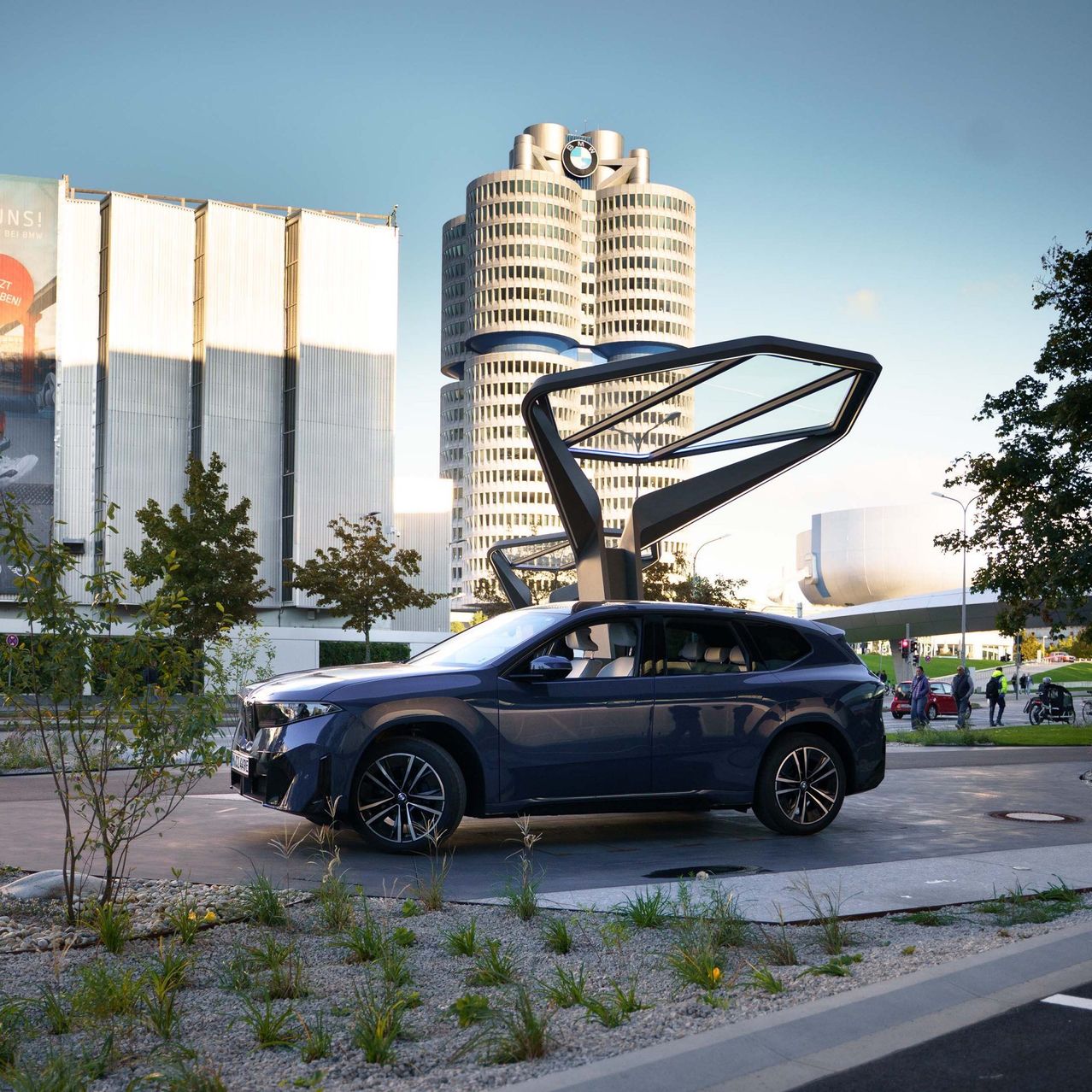 Ein blauer BMW SUV vor einem modernen Gebäude mit großem BMW-Logo, umgeben von Bäumen und urbaner Umgebung.