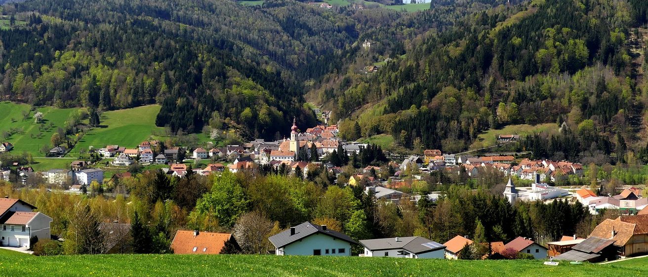 Ein malerisches Dorf in den Bergen, umgeben von üppigem Grün und Häusern mit roten Dächern. In der Mitte steht eine Kirche mit einem Turm.