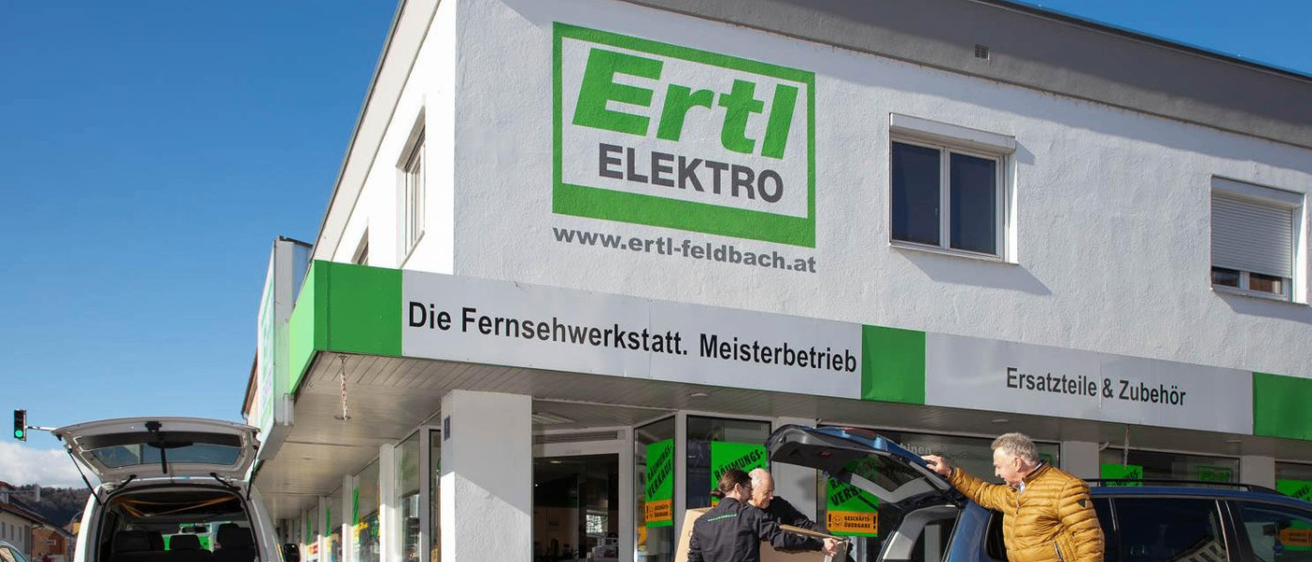 Zwei Männer entladen Kisten aus dem Kofferraum eines blauen Autos vor einem Geschäft namens Ertl Elektro. Das Geschäft zeigt ein grün-weißes Logo, eine Website-Adresse und ein Schild, das 'Die Fernsehwerkstatt. Meisterbetrieb' besagt.