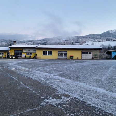 Ein gelbes Gebäude mit einem Schild, das 'SantEr' liest, steht vor einer verschneiten Landschaft. Das Gebäude hat zwei Garagen, und ein Auto ist davor geparkt. Schnee bedeckt den Boden und Berge sind im Hintergrund sichtbar.