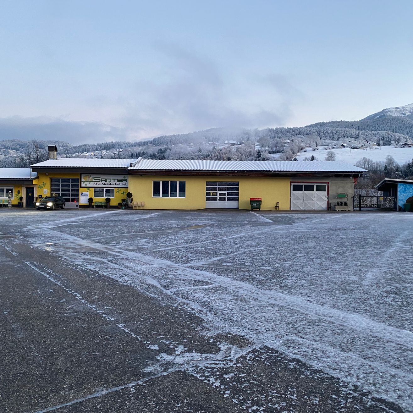 Ein gelbes Gebäude mit einem Schild, das 'SantEr' liest, steht vor einer verschneiten Landschaft. Das Gebäude hat zwei Garagen, und ein Auto ist davor geparkt. Schnee bedeckt den Boden und Berge sind im Hintergrund sichtbar.