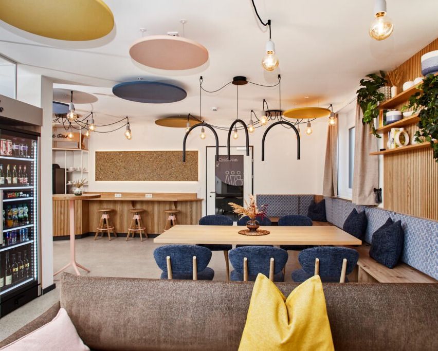 Ein Innencafe mit modernen Möbeln, Holztheken und Hockern. Der Raum hat einen Tisch, Stühle und ein Sofa. Die Decke ist mit verschiedenen Deckenleuchten geschmückt.