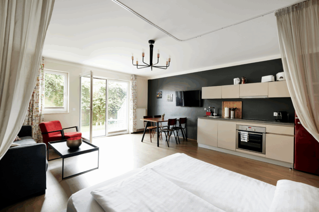 Ein modernes Apartment-Interieur mit Wohnbereich, Esstisch, Küche und Schlafzimmer. Der Raum zeichnet sich durch Holzböden, weiße Wände und eine graue Akzentwand aus.
