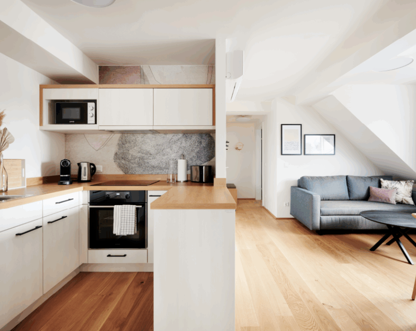 Eine moderne Küche mit Holzboden, weißen Schränken und einer kleinen Mikrowelle. In der Nähe ein graues Sofa und ein Couchtisch. Die Wände sind weiß und es hängen gerahmte Bilder an der Wand.