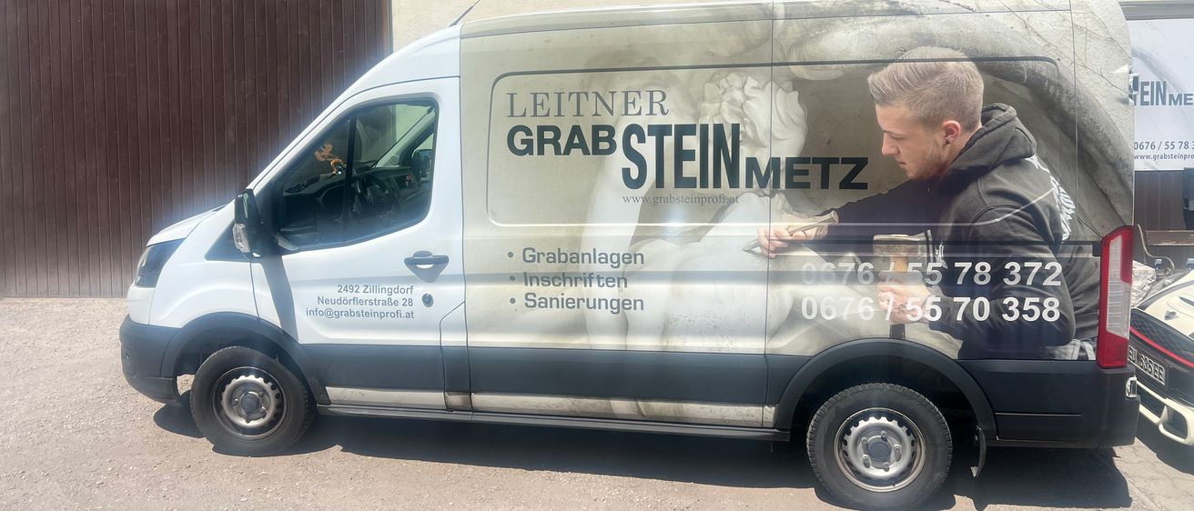 Ein weißer Lieferwagen, der am Straßenrand geparkt ist, mit einem Mann, der etwas daran repariert. Es hat die Worte 'Leitner Grabsteinmetz' und eine Telefonnummer auf der Seite.