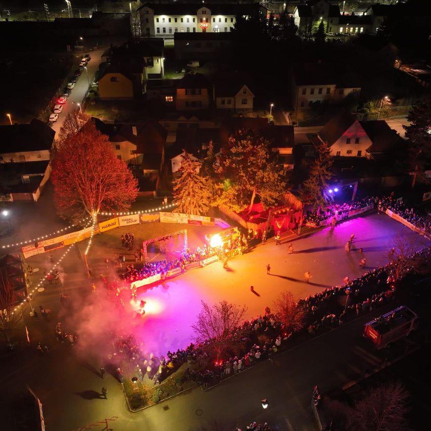 Eine Nachtaufnahme aus der Luft von einer lebhaften Veranstaltung im Freien. Der Veranstaltungsbereich ist mit bunten Lichtern und Rauch beleuchtet. Menschen versammeln sich um den Veranstaltungsbereich und beobachten die Aktivität. Im Hintergrund sind Gebäude und Bäume zu sehen.
