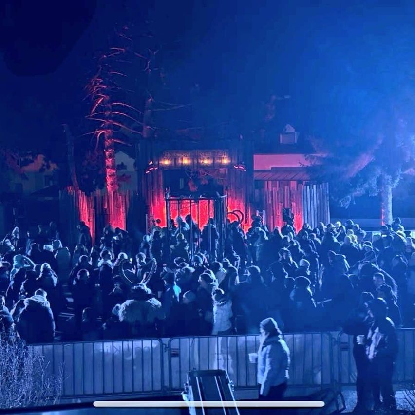 Eine Menschenmenge versammelt sich bei einer nächtlichen Veranstaltung mit einer von roten Lichtern beleuchteten Bühne. Bäume und ein Gebäude sind im Hintergrund zu sehen. Menschen stehen hinter einem Zaun.