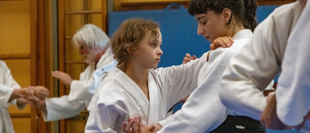 Zwei junge Mädchen in weißen Kampfsportuniformen üben eine Technik, wobei ein Mädchen den Arm des anderen festhält.