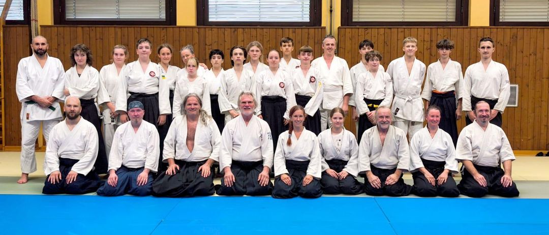 Eine Gruppe von Kampfsportlern in weißen Uniformen und schwarzen Gürteln posiert für ein Foto in einem Dojo. Sie sitzen auf einem blauen Mattenboden mit hölzernen Wänden und Fenstern im Hintergrund.