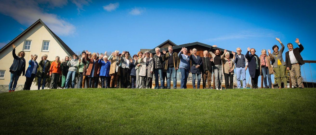 Eine große Gruppe von Menschen steht auf einem Rasen und lächelt und winkt. Sie sind in Winterkleidung gekleidet und scheinen für ein Gruppenfoto zu posieren.