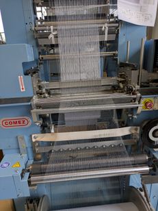 Eine Comez-Maschine zeigt die Verarbeitung eines weißen Fadens, wobei verschiedene Zahnräder und Rollen sichtbar sind. Die Maschine ist blau mit einem gelben Aufkleber auf der linken Seite.