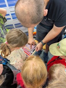 Ein Mann hilft einem jungen Mädchen beim Weben auf einem Webstuhl, umgeben von anderen Kindern, die zuschauen.