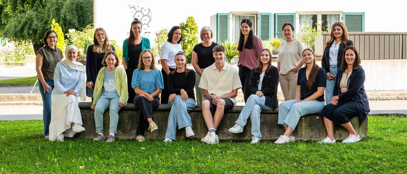 Eine Gruppe von Menschen posiert für ein Foto vor einem weißen Haus. Einige sitzen auf einer Steinbank, andere stehen dahinter. Bäume und Pflanzen umgeben das Haus.
