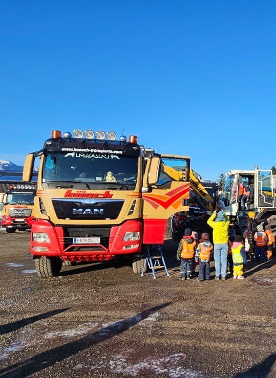 Eine Gruppe von Kindern und Erwachsenen steht um einen gelben und roten MAN-Lkw mit einer großen Kabine herum, unter einem klaren blauen Himmel, in der Nähe eines Gebäudes und anderer Lastwagen.