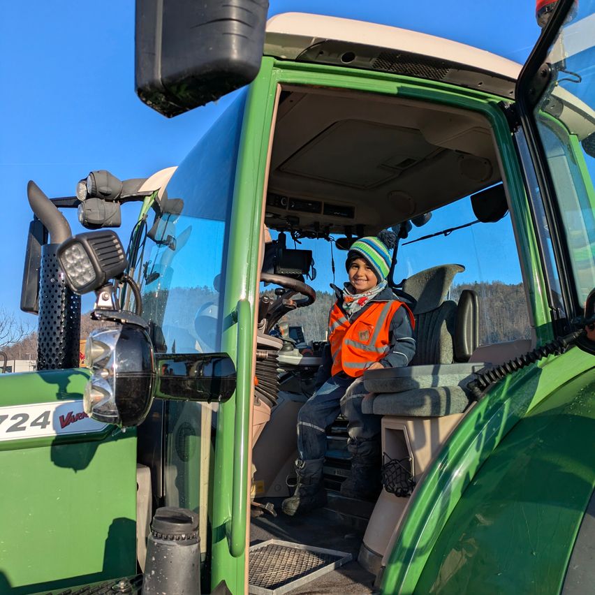 Ein junger Junge in einer Sicherheitsweste sitzt in der Kabine eines grünen Traktors mit der Nummer 724 an der Seite.