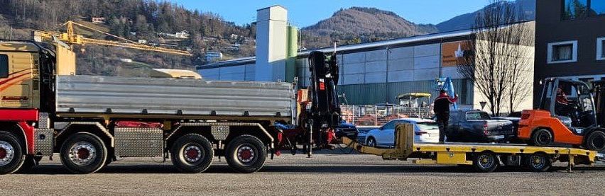 Ein Lkw mit einem Tieflader entlädt Fracht auf einem Parkplatz mit mehreren Autos und einem Gebäude im Hintergrund.