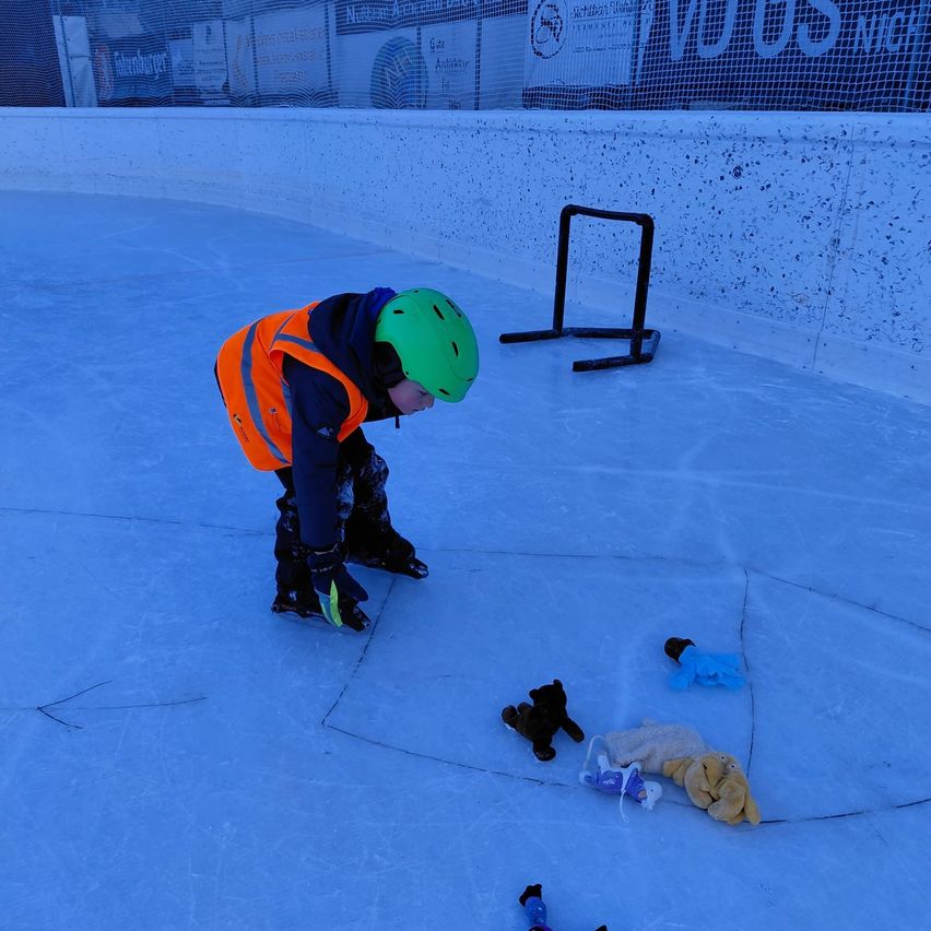 A child wearing a green helmet and an orange vest is bending over on the ice rink, possibly picking up a stuffed toy.