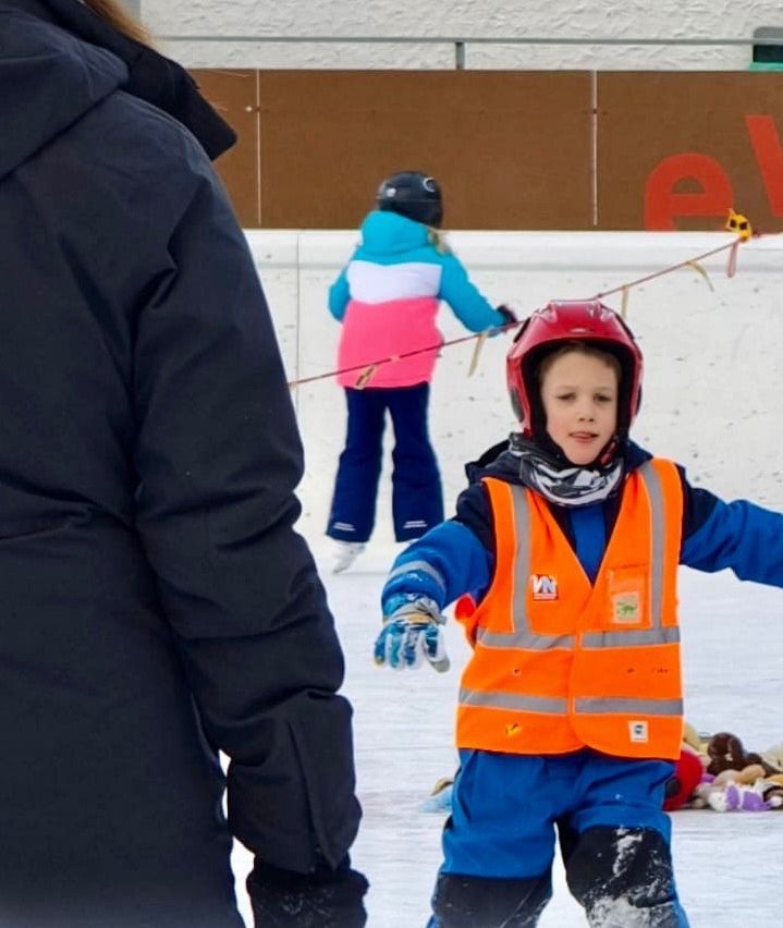 Two children are on an ice rink, one wearing a reflective vest and a helmet, the other in a blue and pink jacket, both holding onto ropes. A person in a black jacket is partially visible.