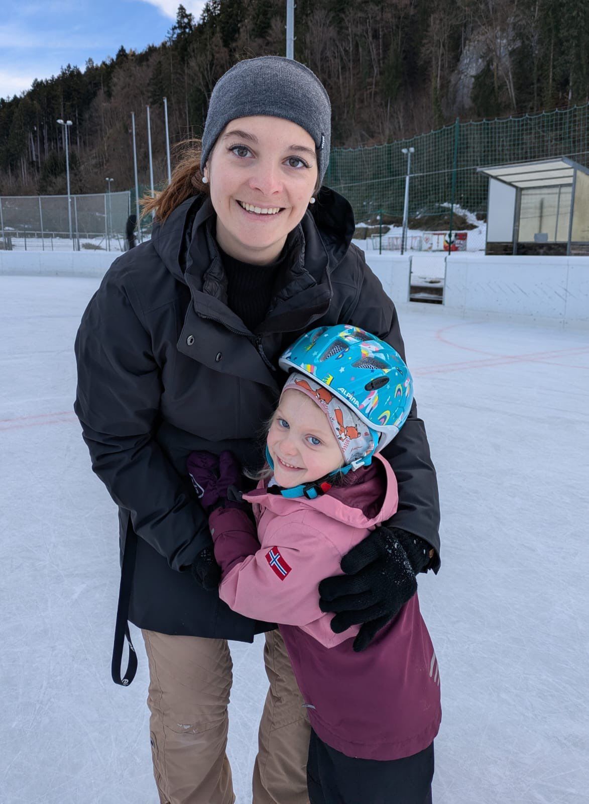 A woman with a smiling face stands on an ice rink and holds a child in her arms. The child is wearing a helmet and gloves.