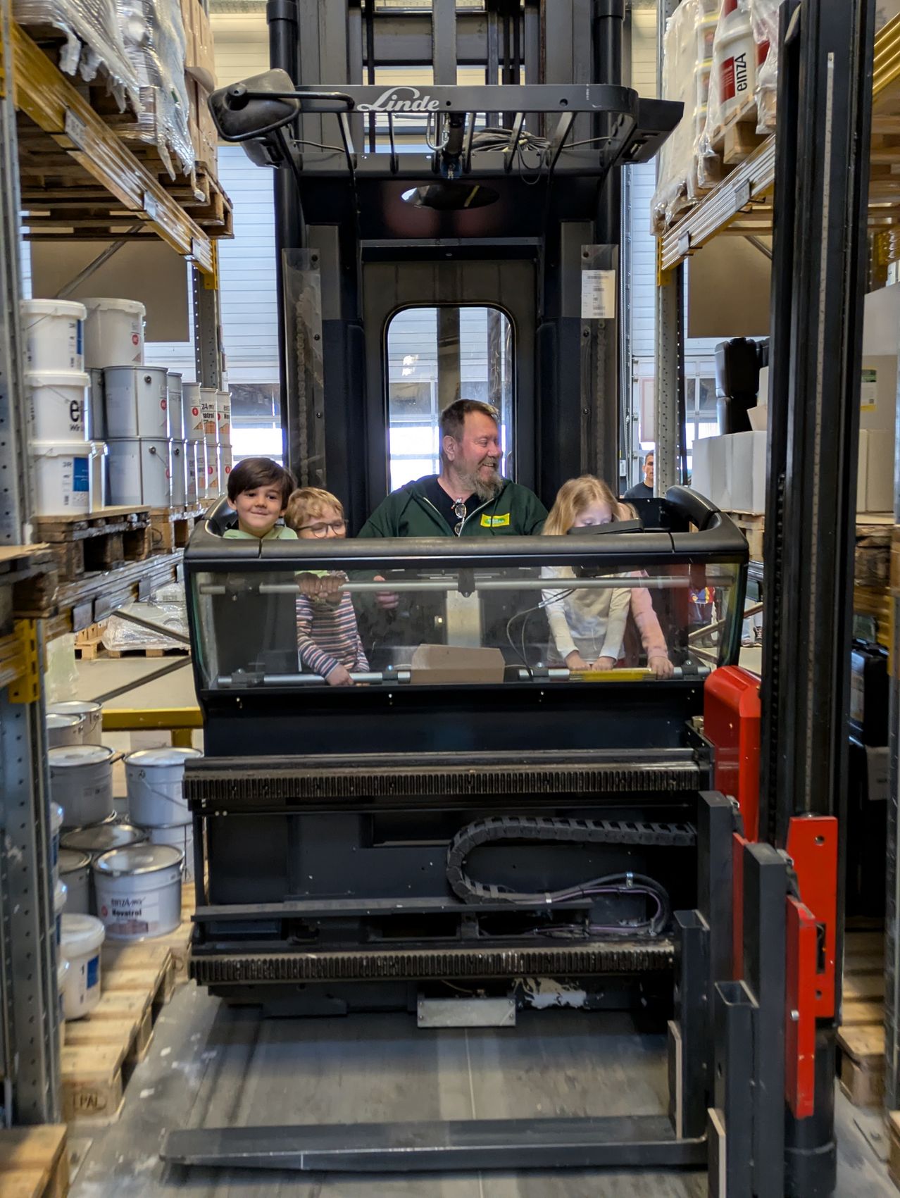 Ein Mann mit drei Kindern sitzt in einem Gabelstapler in einem Lagerhaus. Der Mann lächelt und die Kinder sehen neugierig aus. Der Gabelstapler steht auf dem Boden und es gibt große Container dahinter.