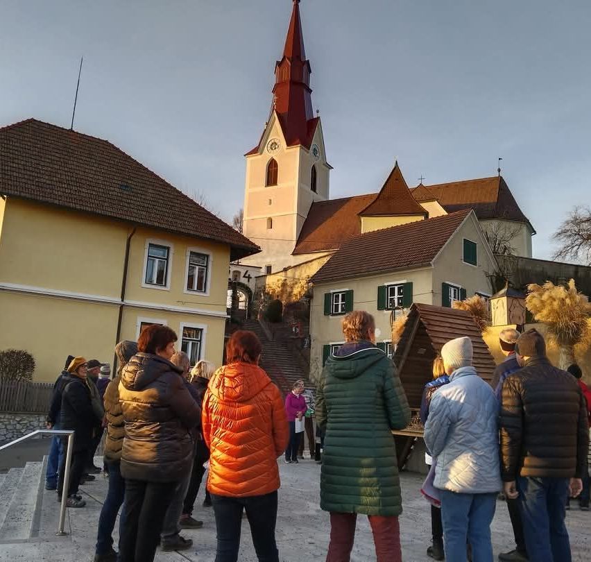 Eine Gruppe von Menschen steht vor einer Kirche in einem Dorf mit Turm und Dachreiter unter klarem Himmel.