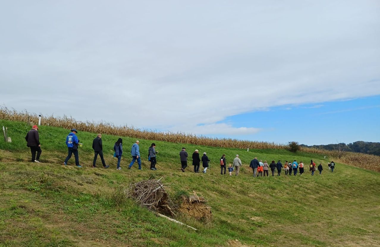 Eine Gruppe von Menschen wandert auf einem grasbewachsenen Hügel mit einem Maisfeld im Hintergrund. Sie sind in lässiger Outdoor-Kleidung gekleidet.