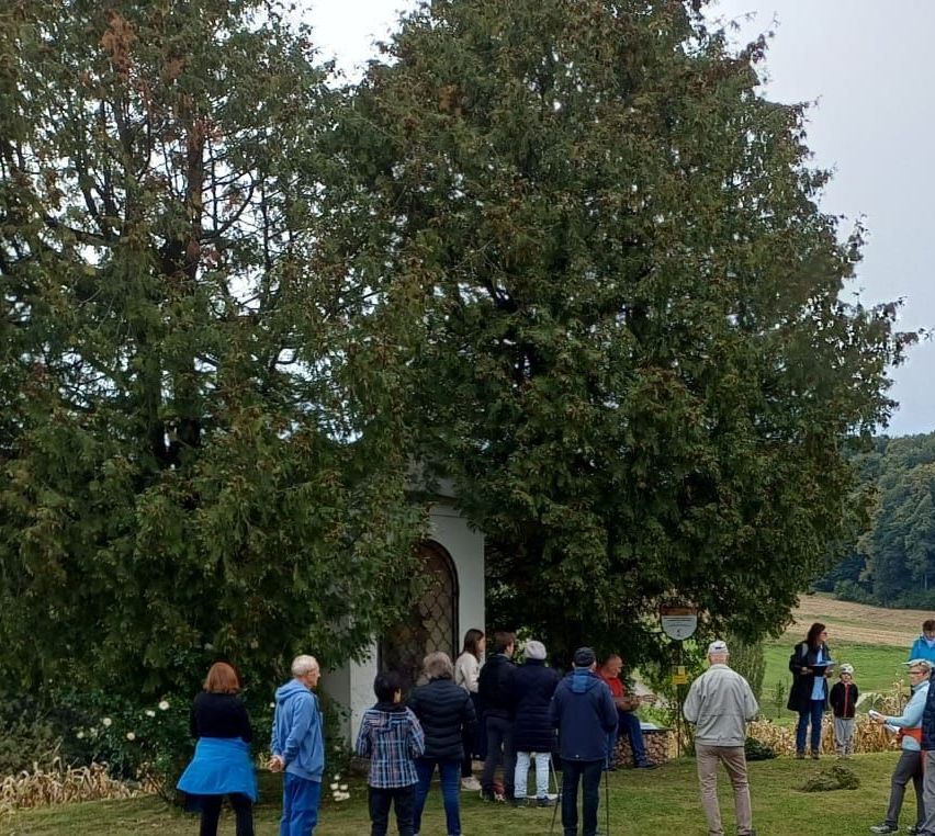 Eine Gruppe von Menschen steht auf einem Feld mit einem großen Baum und einem kleinen Gebäude. Einige sind in Jacken und Mützen gekleidet, andere tragen Mützen. Eine Frau spricht zu ihnen.