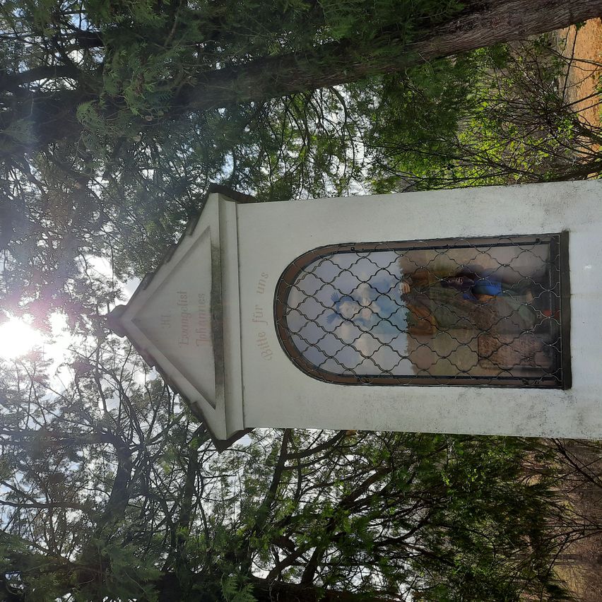 A white chapel with a glass window showing a religious figure under a bright sun, surrounded by trees.
