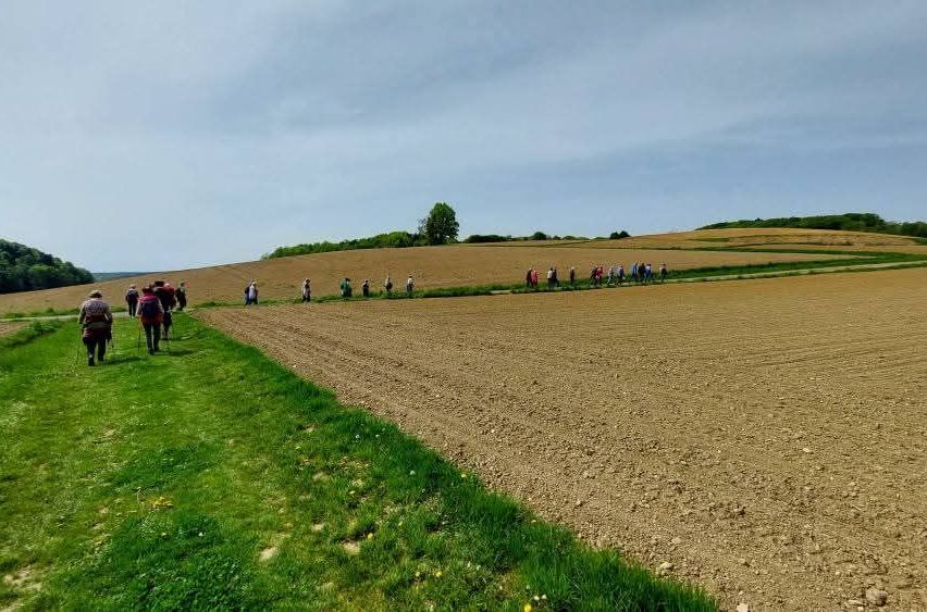 A group of people are hiking on a grassy path alongside a vast, barren field under a partly cloudy sky.
