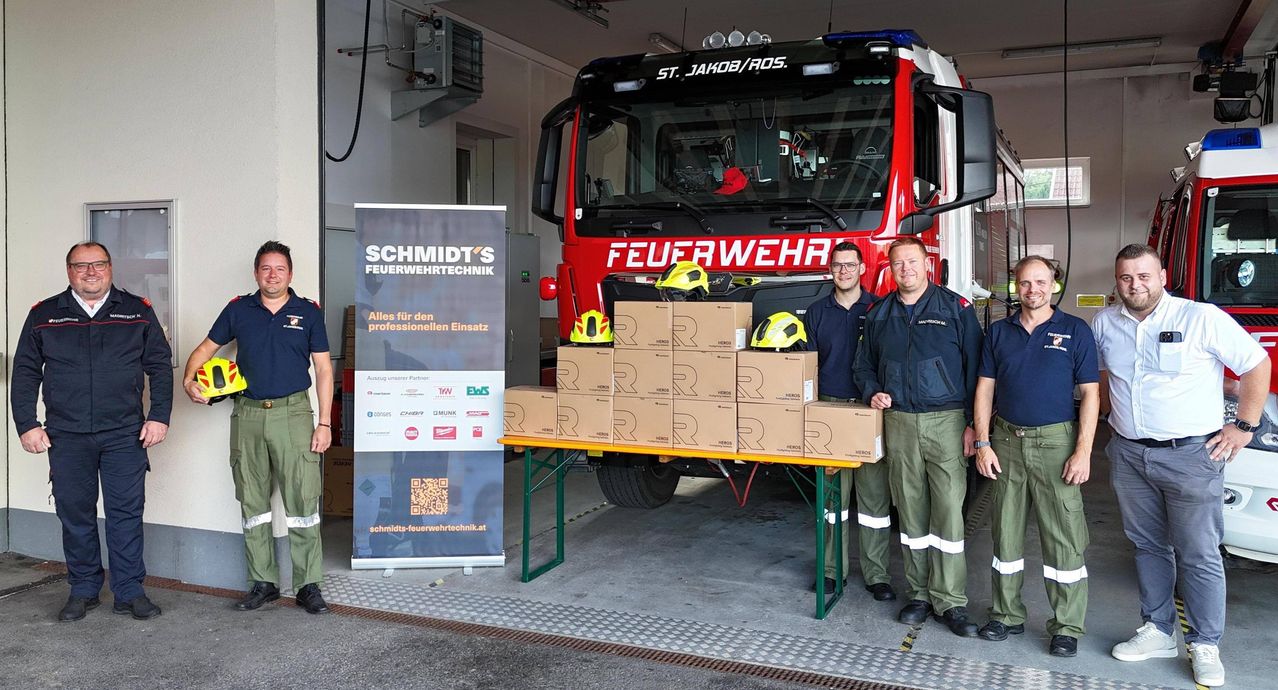 Drei Männer stehen lächelnd neben einem Feuerwehrauto. Vor ihnen steht ein Tisch mit Kisten. Ein Banner mit dem Text SCHMIDT'S FEURWEHRTECHNIK ist links.
