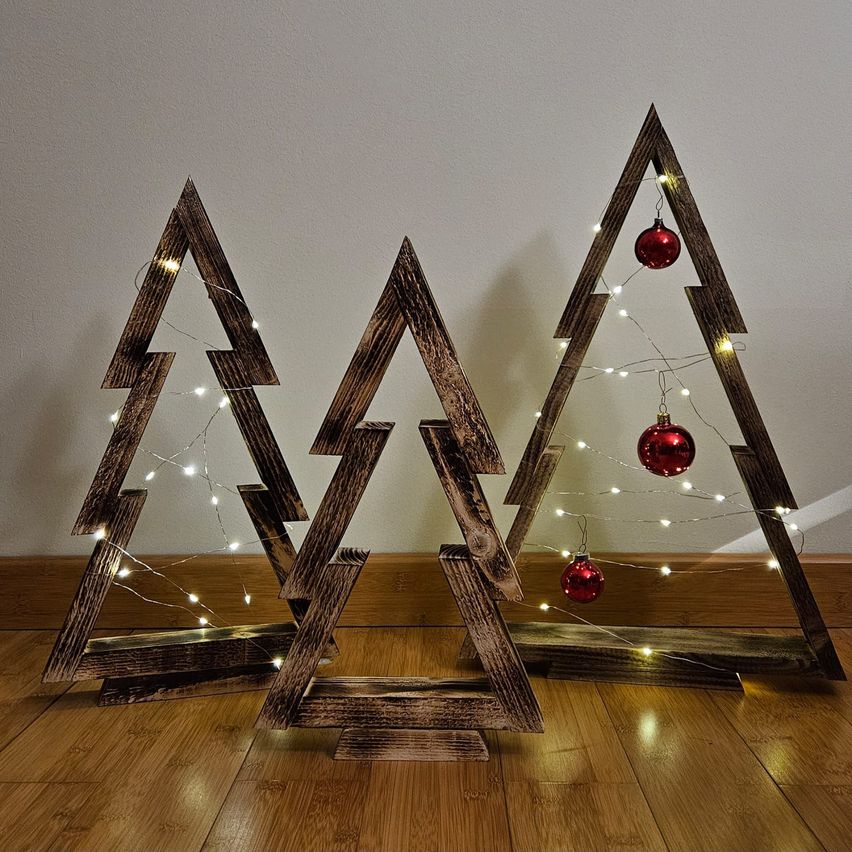 Drei rustikale Holz-Weihnachtsbäume mit Lichterketten und roten Kugeln sind auf einem Holzboden angeordnet. Die Bäume sind dreieckig und haben einen warmen, rustikalen Look.