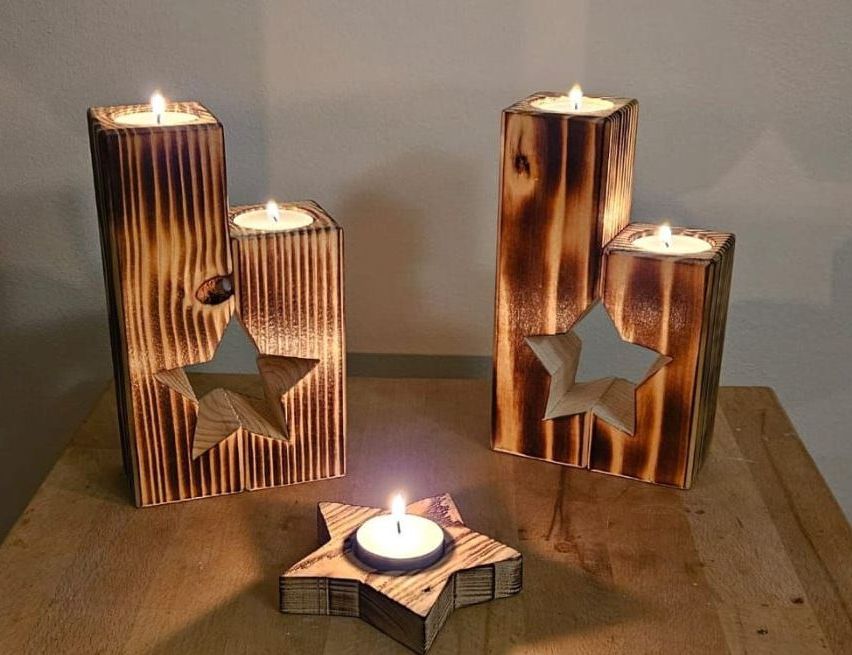 Drei hölzerne Kerzenhalter mit Sternausschnitten sind beleuchtet. Zwei größere Halter haben jeweils zwei Kerzen, während ein kleinerer Halter eine einzelne Kerze hat. Sie stehen auf einem Holztisch.